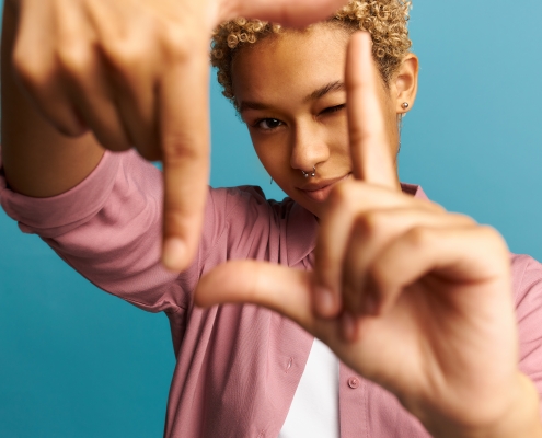Selective focus of smiling black girl with blonde curly hair and piercing making frame with hands and fingers, looking through it, closing one eye. Creativity and photography concept. Body language