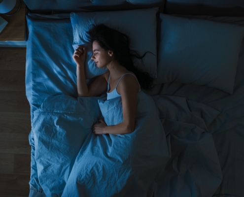 Top View of Beautiful Young Woman Sleeping Cozily on a Bed in His Bedroom at Night. Blue Nightly Colors with Cold Weak Lamppost Light Shining Through the Window.